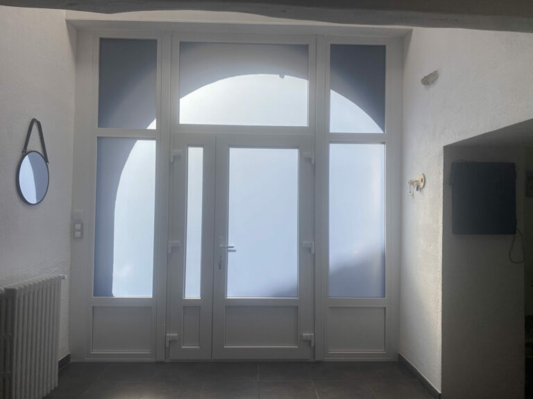 Porte d’entrée vitrée d’immeuble : menuiserie grise recouverte d’un film adhésif blanc, imposte cintrée conservée